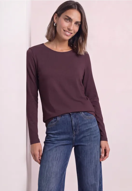 Vrouw in bordeauxrode longsleeve en blauwe spijkerbroek, glimlachend met halflang haar en subtiele oorbellen.