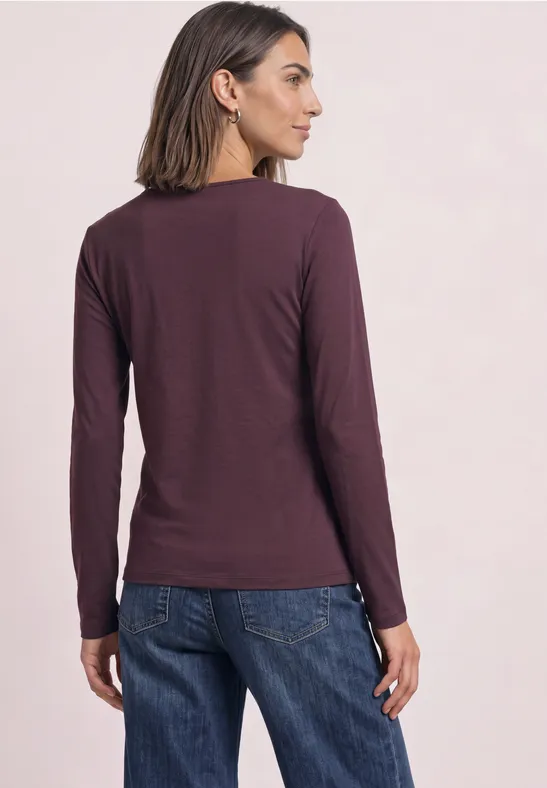 Vrouw van achteren gezien, met een bordeauxrode longsleeve en een spijkerbroek voor een lichte achtergrond.