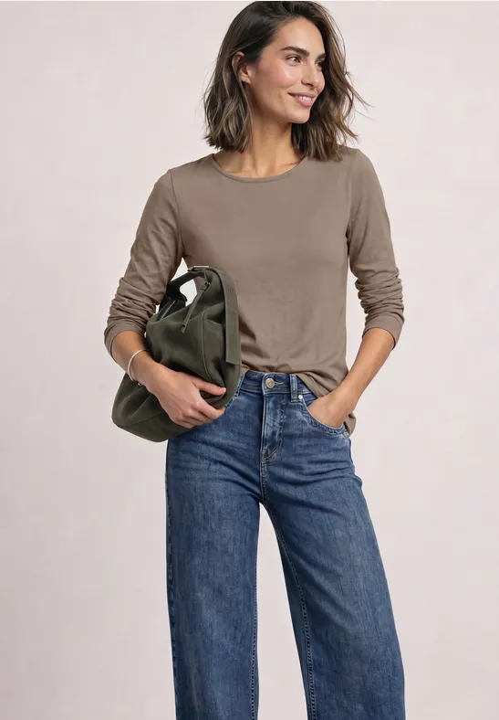 Frau in braunem Langarmshirt, blauen Jeans und grüner Tasche, lächelnd.