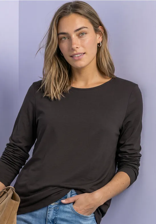 Vrouw in een bruin longsleeve en met tas, nonchalant poseerend voor een lichte achtergrond.