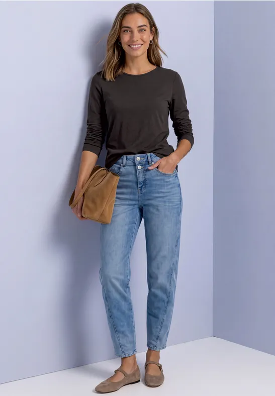 Een vrouw in zwart shirt, blauwe jeans en bruine accessoires staat voor een lichtpaarse muur.