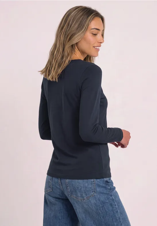 Vrouw in donkerblauw shirt met lange mouwen en spijkerbroek, glimlachend met gedraaid bovenlichaam, lichte achtergrond