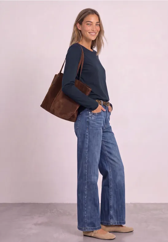Vrouw met blauwe trui en wijde jeans, dragend een bruine tas, glimlachend opzij staand.