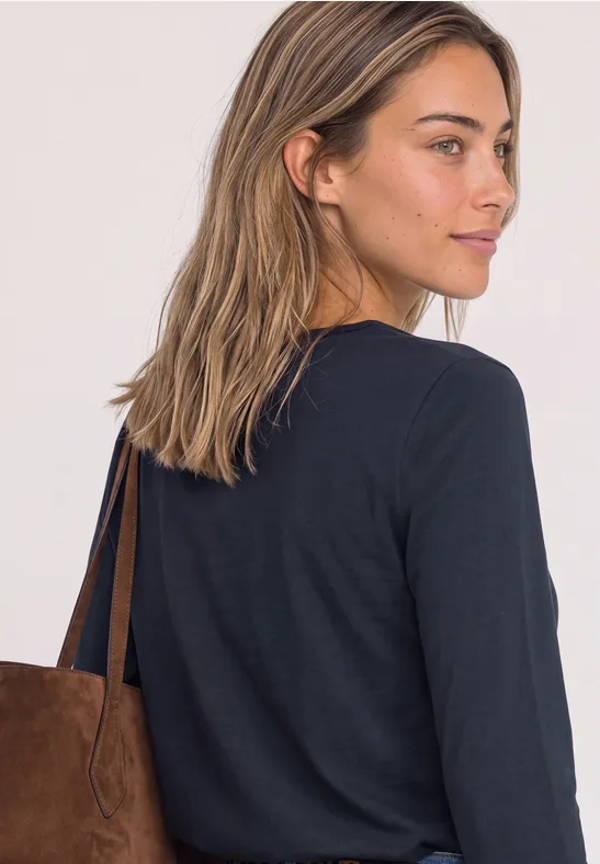 Een vrouw met bruin haar draagt een zwart langarmshirt en een bruine suède tas.
