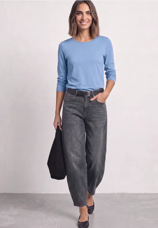 Vrouw in blauw shirt met lange mouwen, losse zwarte spijkerbroek en zwarte schoenen, met tas.