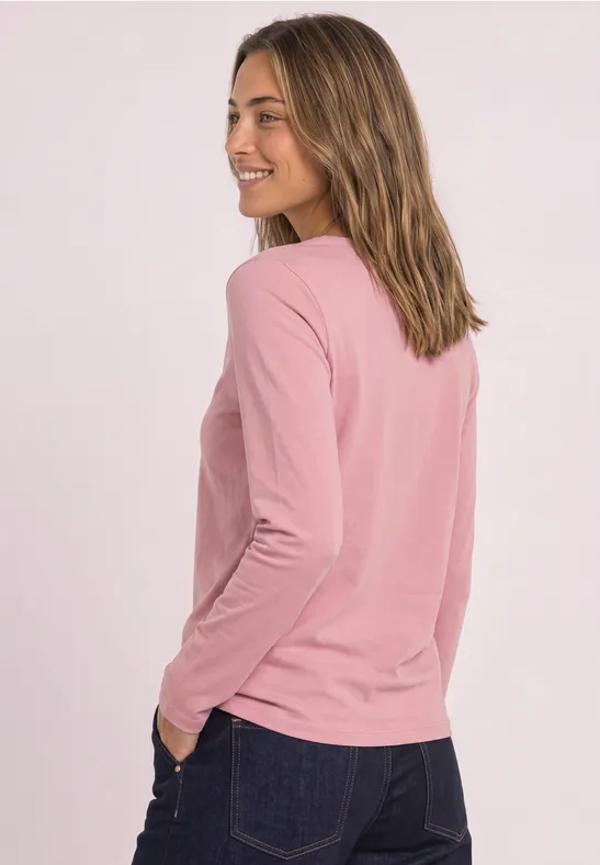 Frau in rosa Langarmshirt und Jeans, lächelnd seitlich stehend vor hellem Hintergrund.