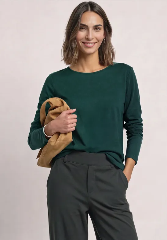 Frau in grünem Longsleeve und dunkler Hose hält eine beige Tasche.