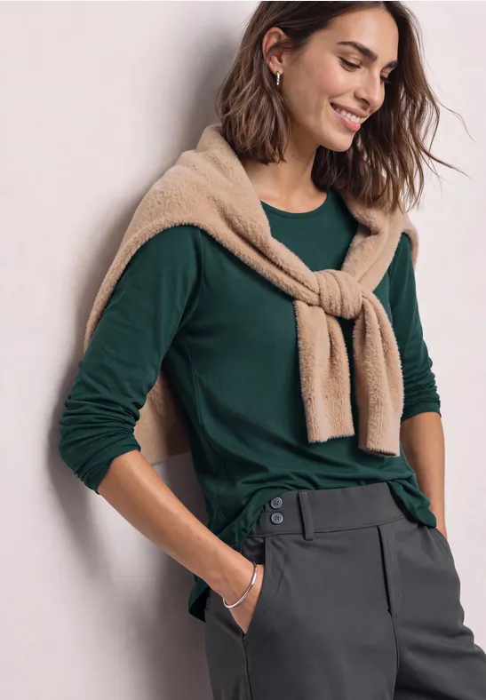 Frau in grünem Langarmshirt mit beiger Strickjacke um die Schultern und grauer Hose, lächelnd.