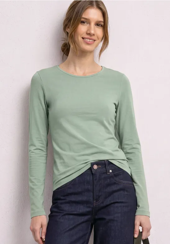 Vrouw in mintgroen longsleeve en donkere jeans, staand lachend voor een lichte achtergrond.