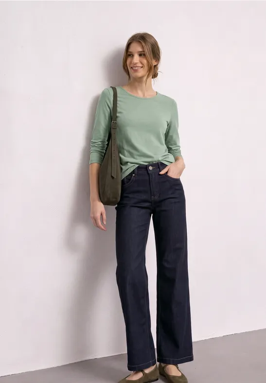 Vrouw met groen shirt, donkerblauwe wijdvallende jeans en schoudertas, staand voor een neutrale muur.