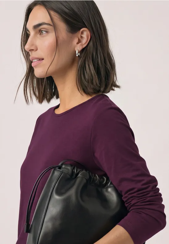 Frau von der Seite in lila Oberteil mit schwarzer Beuteltasche, schulterlangem Haar.