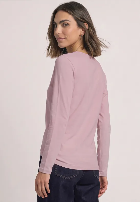Eine Frau in rosa Langarmshirt und dunkler Hose steht seitlich und schaut über die Schulter.