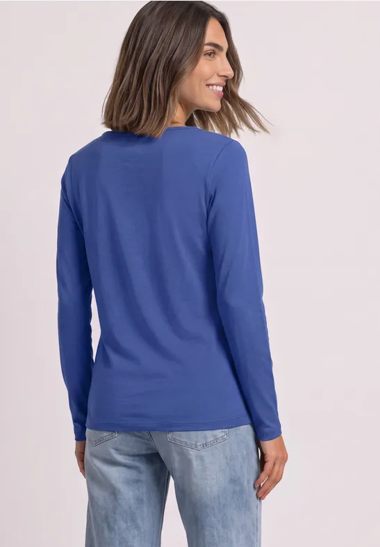 Frau in blauem Langarmshirt und Jeans lächelt, steht seitlich mit Blick über die Schulter.