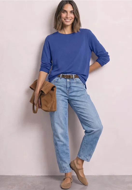 Frau in blauem Pullover und Jeans mit brauner Tasche und Schuhen vor neutralem Hintergrund.