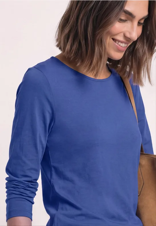 Eine lächelnde Frau in blauem Langarmshirt mit Umhängetasche vor hellem Hintergrund.