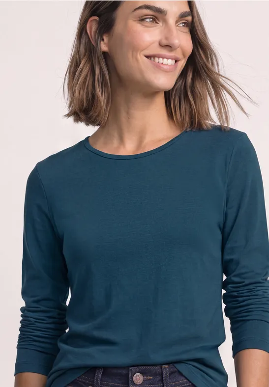 Eine lächelnde Frau trägt ein petrolfarbenes Langarmshirt und Jeans vor hellem Hintergrund.