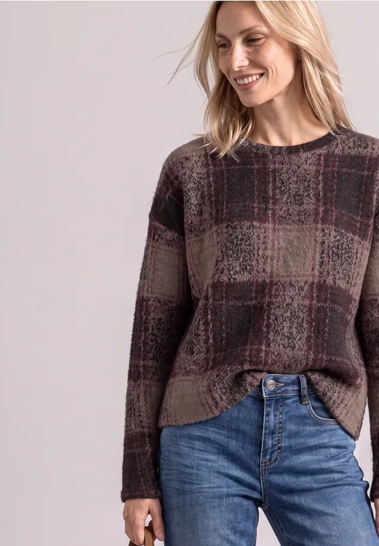 Frau lächelt und trägt einen karrierten Pullover in Erdtönen mit Jeans vor hellem Hintergrund.