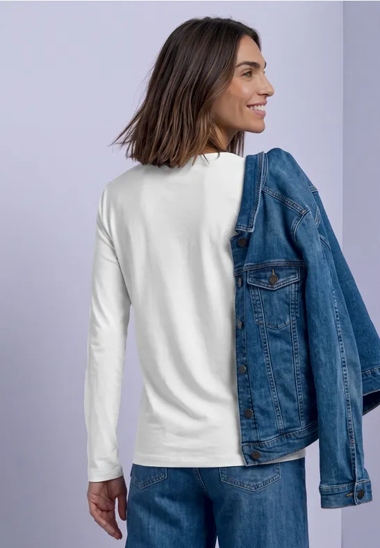 Femme en tee-shirt blanc à manches longues porte une veste en jean sur l'épaule et sourit devant un fond violet clair.