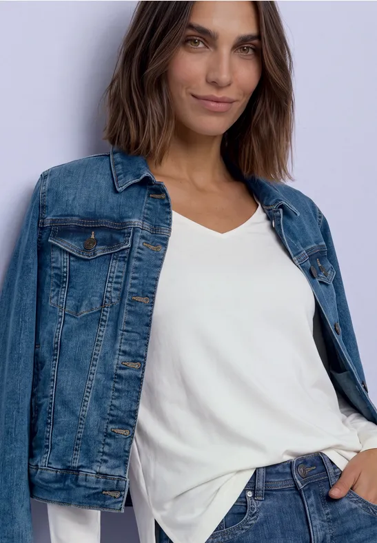 Femme portant une veste en jean et un t-shirt blanc, posant décontractée devant un fond clair.