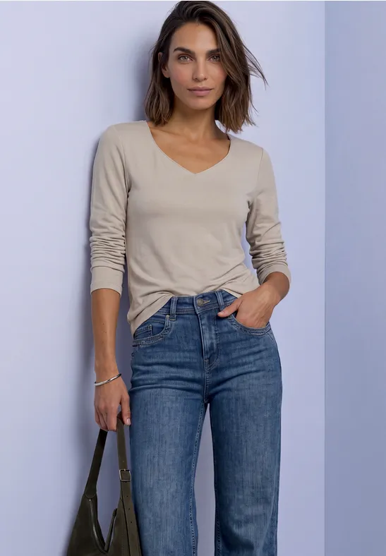 Femme en t-shirt beige à manches longues et jeans bleu, posant décontractée avec sac à bandoulière sur fond uni.