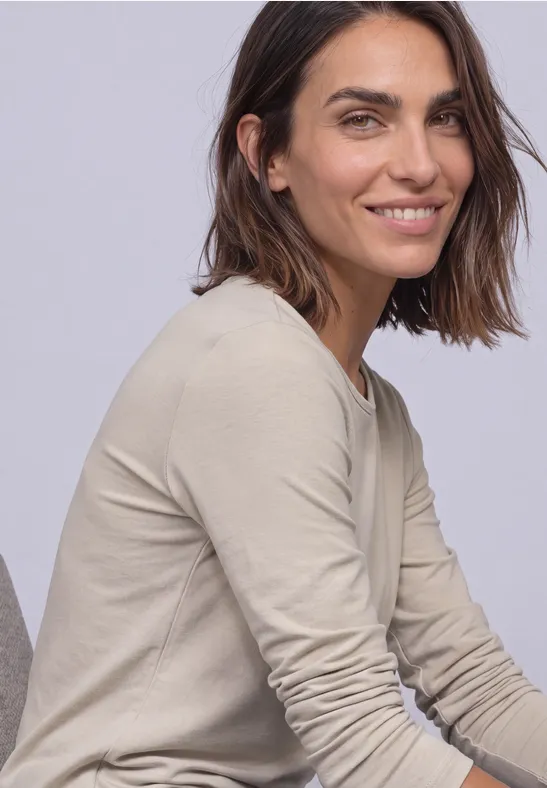 Une femme souriante aux cheveux bruns mi-longs, vêtue d'un haut à manches longues beige sur fond clair.