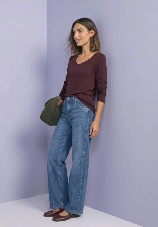 Femme en top bordeaux, jean large bleu, chaussures plates et sac vert devant un mur violet.