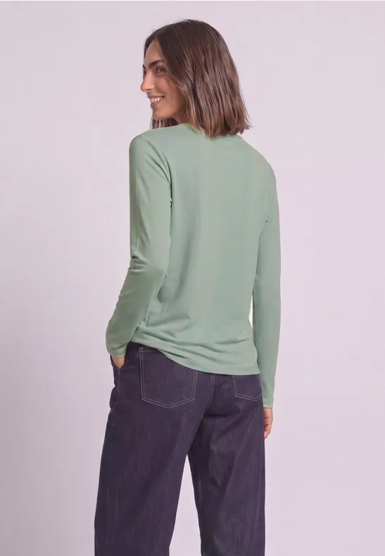 Femme en haut vert à manches longues, de profil, main dans la poche et regard souriant par-dessus l'épaule.