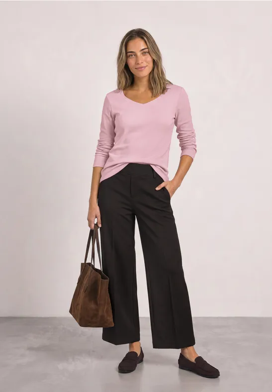 Vrouw in roze V-trui, zwarte wijde broek, bruine tas en zwarte mocassins voor lichte achtergrond.