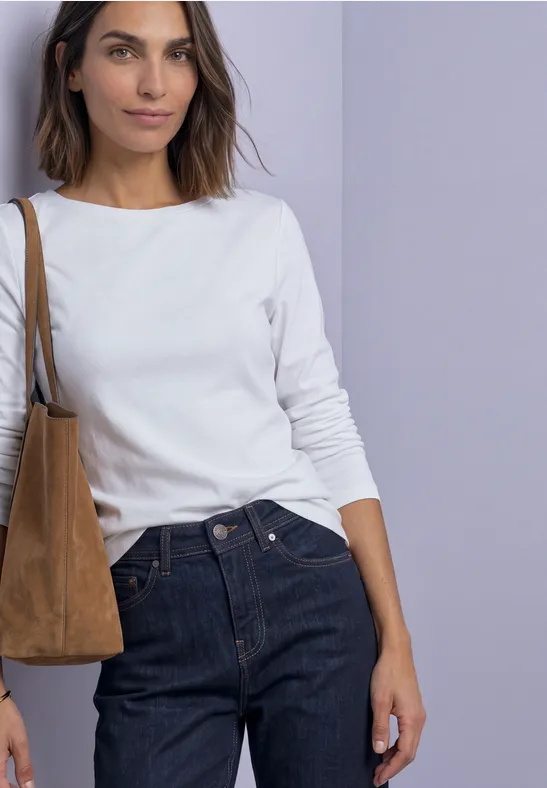 Vrouw in wit shirt met lange mouwen, donkerblauwe jeans en bruine suède tas voor een lichte achtergrond.