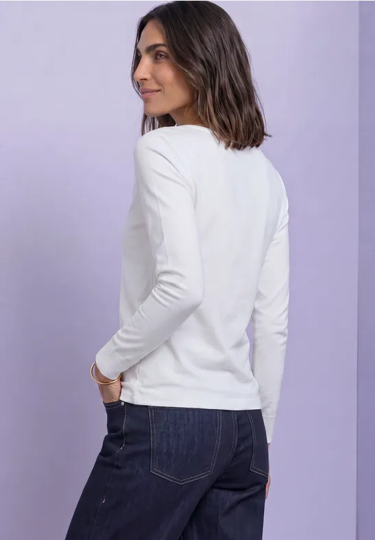 Vrouw in wit lang shirt en spijkerbroek, zijaanzicht tegen zachtpaarse achtergrond.