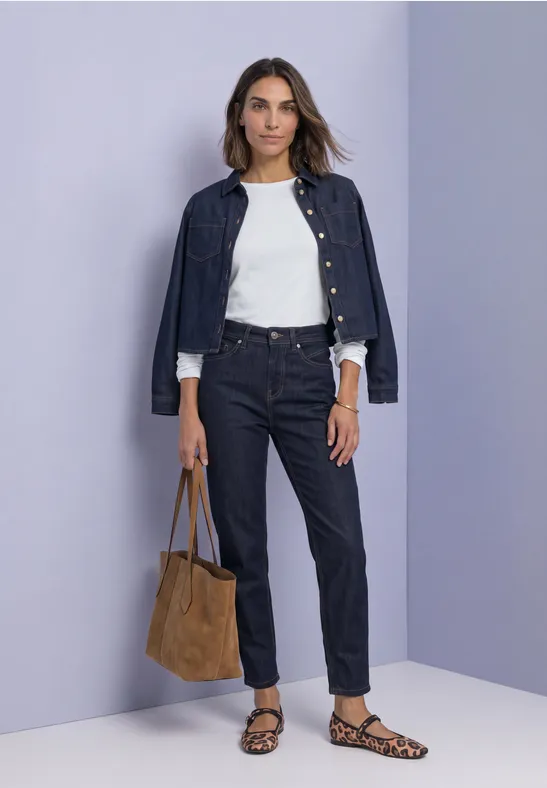 Vrouw met donker denimjasje en -jeans, wit t-shirt, luipaardsandalen en bruine tas tegen paarse achtergrond.
