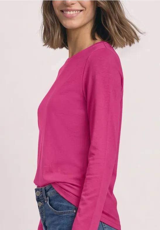 Frau im pinken Langarmshirt und Jeans, seitlich stehend, lächelnd vor hellem Hintergrund.