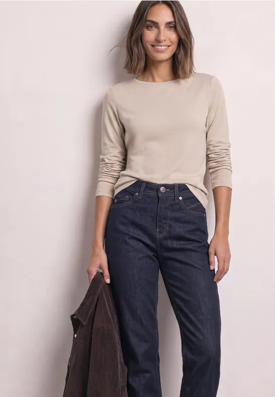 Frau mit beigeem Langarmshirt und hoher Jeans, hält dunklen Mantel in einer Hand, lächelt vor hellem Hintergrund.