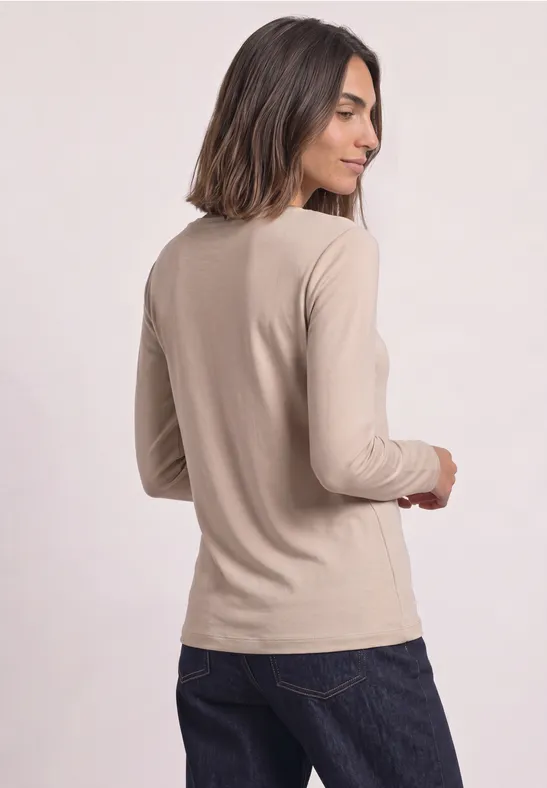 Frau mit schulterlangen dunklen Haaren trägt beige Langarmshirt und dunkle Jeans vor hellem Hintergrund.