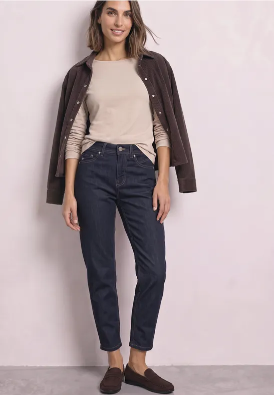 Frau mit beiger Bluse, dunkler Lederjacke über den Schultern, blauen Jeans und braunen Schuhen vor rosafarbenem Hintergrund.