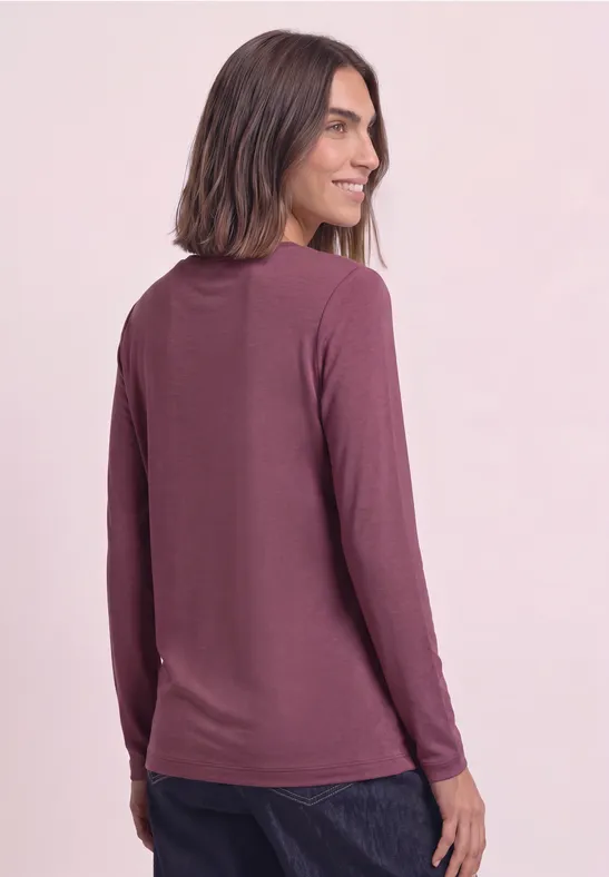 Frau mit schulterlangem Haar in lässigem langärmligem Shirt in Mauve, schaut über die Schulter und lächelt.