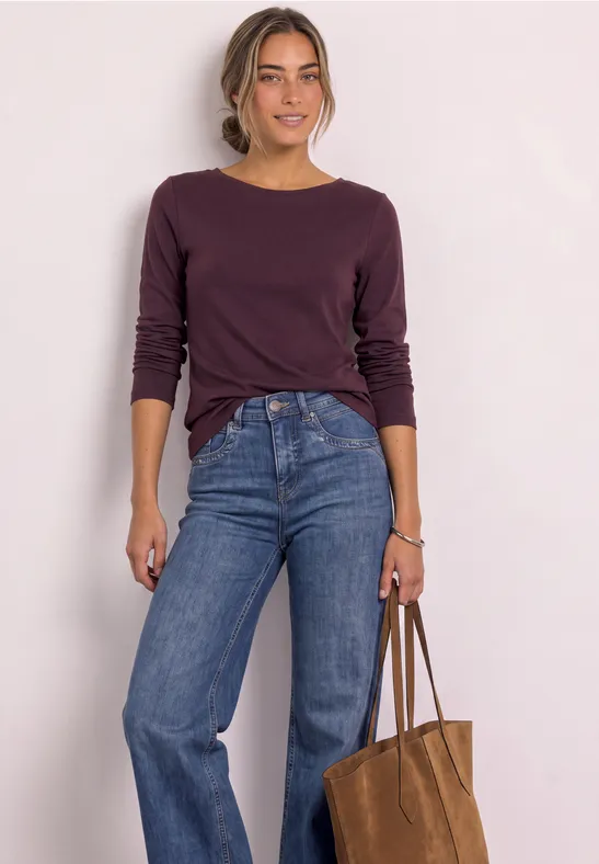 Frau vor hellem Hintergrund in lila Langarmshirt, blauen Jeans und brauner Tasche.