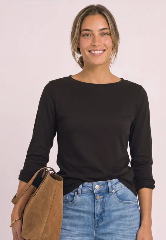 Eine lächelnde Frau in schwarzem Langarmshirt und Jeans hält eine braune Wildledertasche vor hellem Hintergrund.