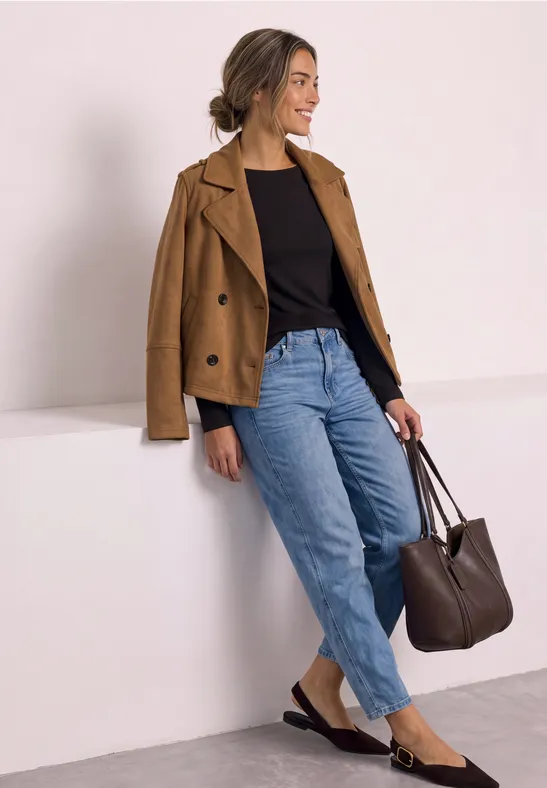 Eine Frau in lässiger Kleidung mit brauner Jacke, schwarzem Shirt, Jeans und Tasche lehnt an einer weißen Wand.
