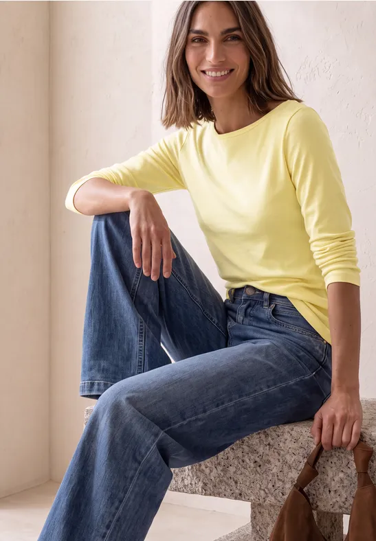 Vrouw zit lachend op een blok, draagt een geel shirt, blauwe jeans en houdt een bruine tas vast.