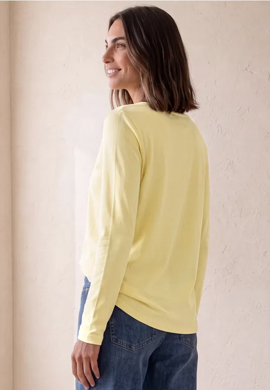 Vrouw in geel shirt met lange mouwen, zijdelings pose, kijkt naar links tegen lichte achtergrond.