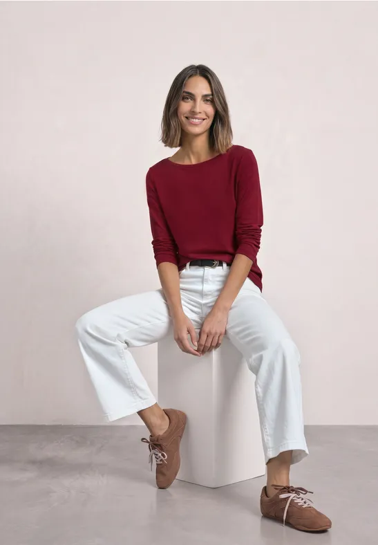 Femme souriante en pull bordeaux, pantalon blanc et mocassins bruns, assise sur un cube blanc.