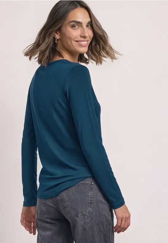 Vrouw in blauw shirt met lange mouwen en donkere jeans glimlacht en kijkt schuin over haar schouder.