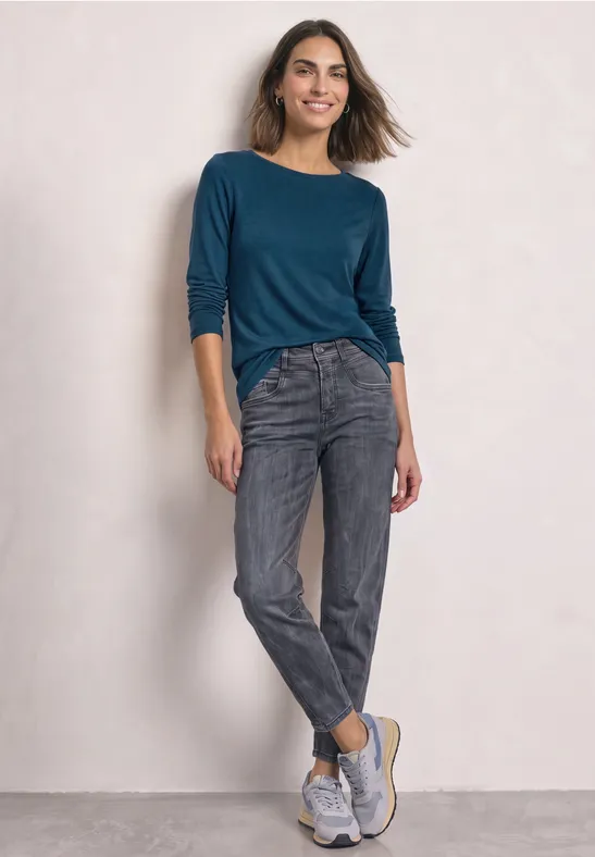Een vrouw draagt een blauwgroen shirt, grijze jeans en witte sneakers tegen een lichte achtergrond.