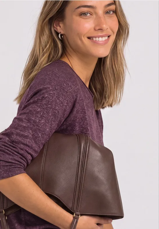 Eine lächelnde Frau in lila Pullover hält eine braune, trapezförmige Ledertasche seitlich unter dem Arm.