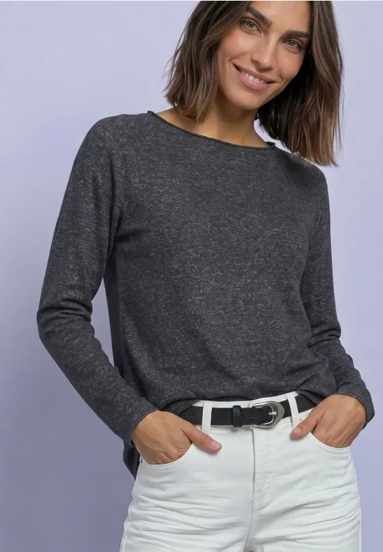 Frau im dunkelgrauen Langarmshirt, weißen Hosen mit Gürtel, lächelnd vor hellem Hintergrund.