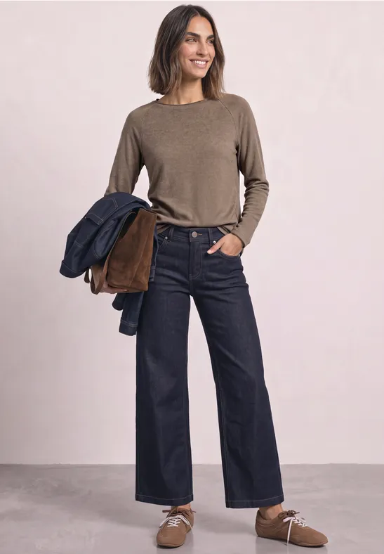 Frau in braunem Pullover und weitem Jeans hält Jacken auf dem Arm.