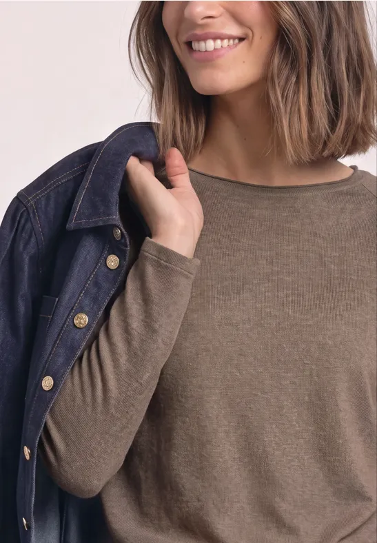 Frau in braunem Langarmshirt hält lässig eine Jeansjacke über die Schulter und lächelt.