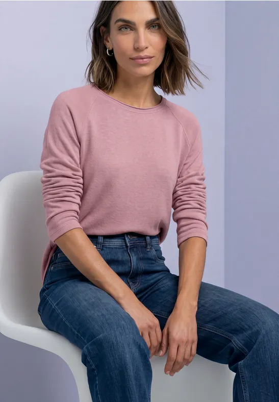 Eine Frau sitzt auf einem weißen Stuhl, trägt einen rosa Pullover und blaue Jeans.