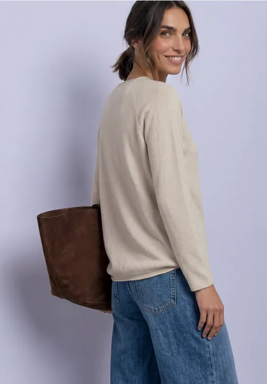 Eine Frau mit beigem Pullover, Jeans und brauner Tasche blickt lächelnd über die Schulter.